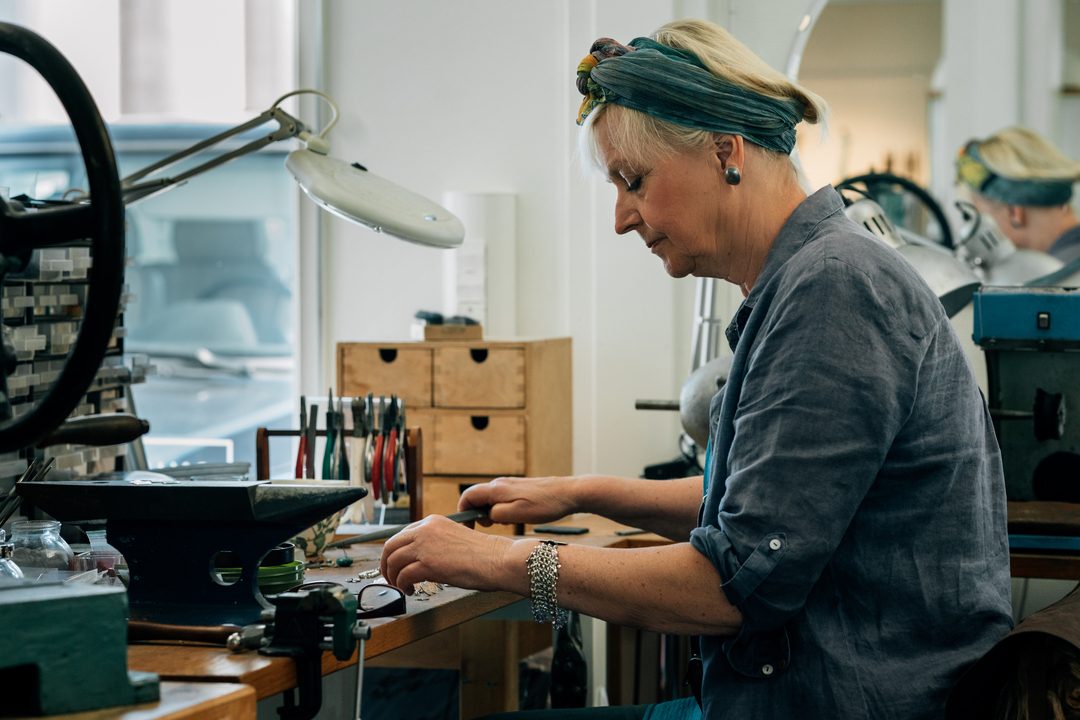 En kvinna sitter vid ett arbetsbord i en verkstad och arbetar koncentrerat med små verktyg. Hon har en grön huvudduk, kort ljust hår och bär en blå skjorta. På bordet finns smyckesverktyg, en liten städ, lådor och en arbetslampa, vilket antyder att hon arbetar med smyckestillverkning eller finmekanik i en ljus och ordnad arbetsmiljö. Kvinnan jobbar på Slemmestad centrum i Norge.