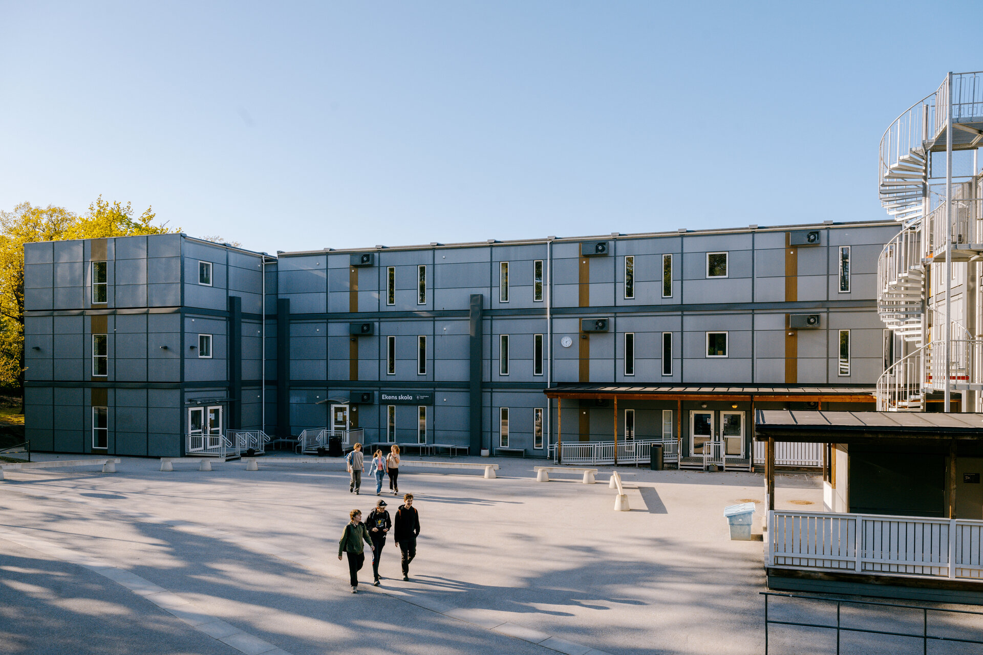 Ekens skola i Hammarby Sjöstad med en skolmodullösning från Expandia, där elever rör sig över skolgården framför grå moduler i två plan med entréer och utvändiga trappor.