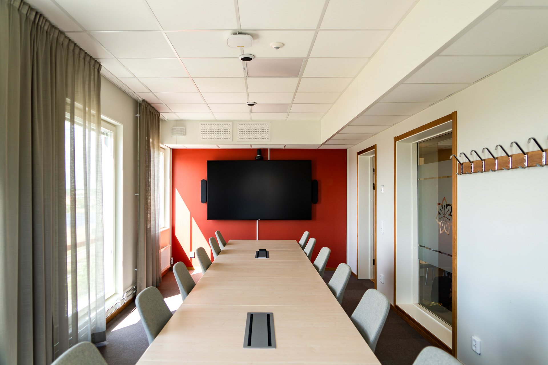 Ett modernt konferensrum med ett långt mötesbord och stolar på båda sidor, en stor skärm monterad på en röd fondvägg och generöst ljusinsläpp från fönster med gardiner. Rummet ingår i kontorsmoduler från Expandia vid Centralsjukhuset Karlstad och är anpassat för digitala möten, presentationer och effektivt samarbete.