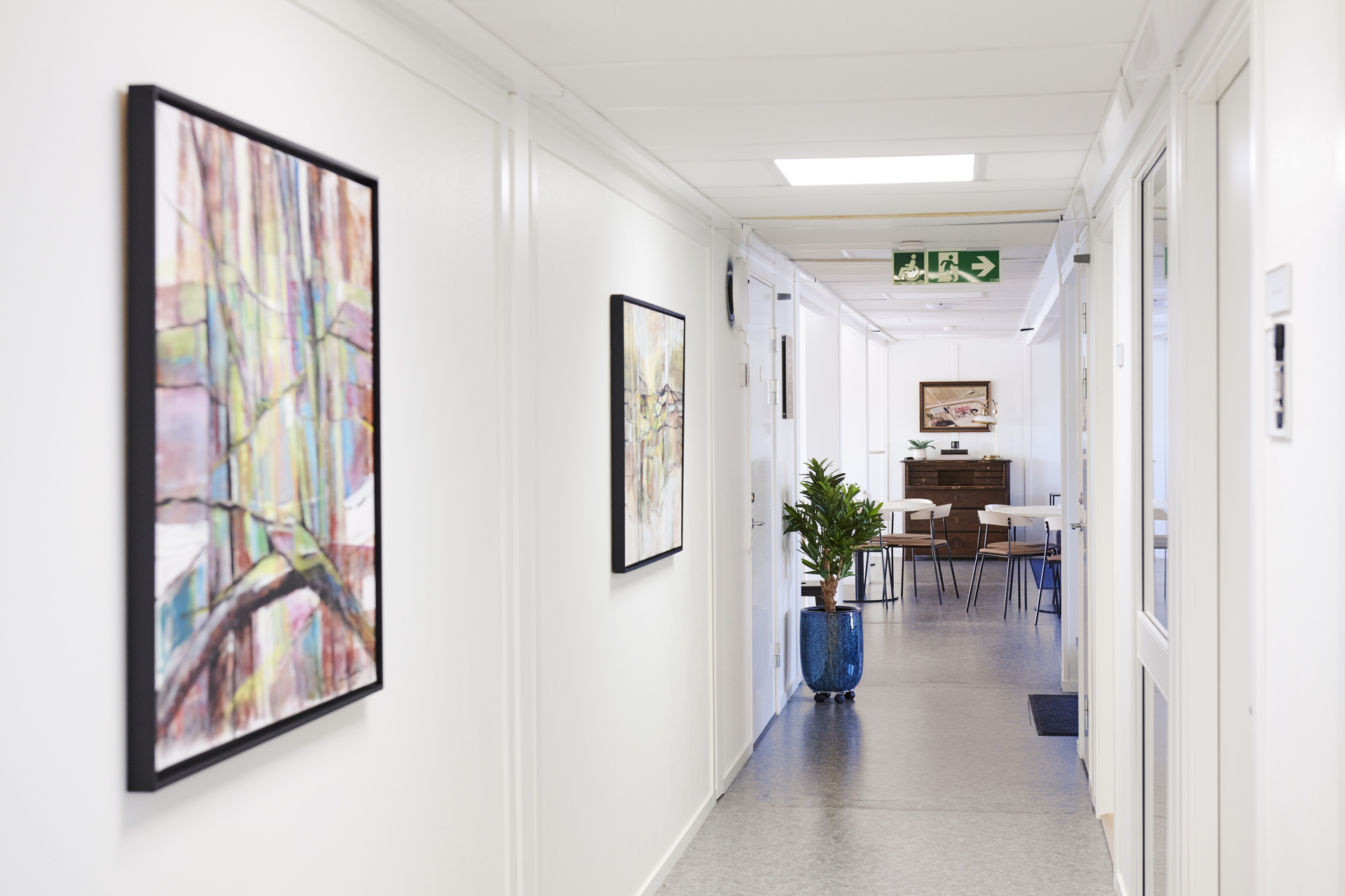 Ljus korridor i kontorsmoduler från Expandia med konst på väggarna, dörrar till kontorsrum och en växt i kruka. I bakgrunden syns ett gemensamt utrymme med bord och stolar. Bilden är tagen i kontorsmoduler vid Alviks trafik i Luleå.