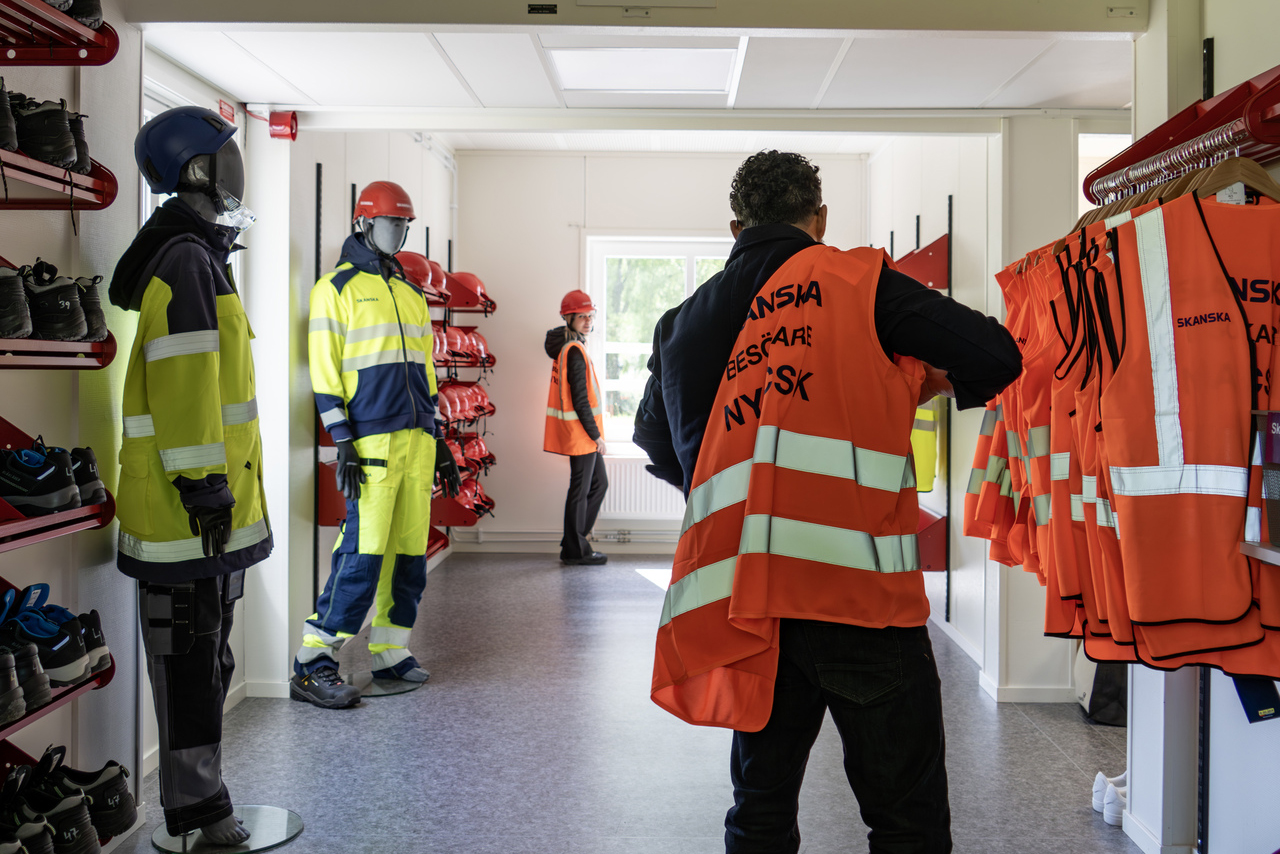 Utrymme för skyddsutrustning i kontorsmoduler från Expandia i Skanskas projektkontor i Karlstad. En person tar på sig en varselväst, medan skyddshjälmar, kläder och skor är organiserade på hyllor och ställ längs väggarna. Rummet är ljust och anpassat för säkerhetsgenomgång och ombyte.