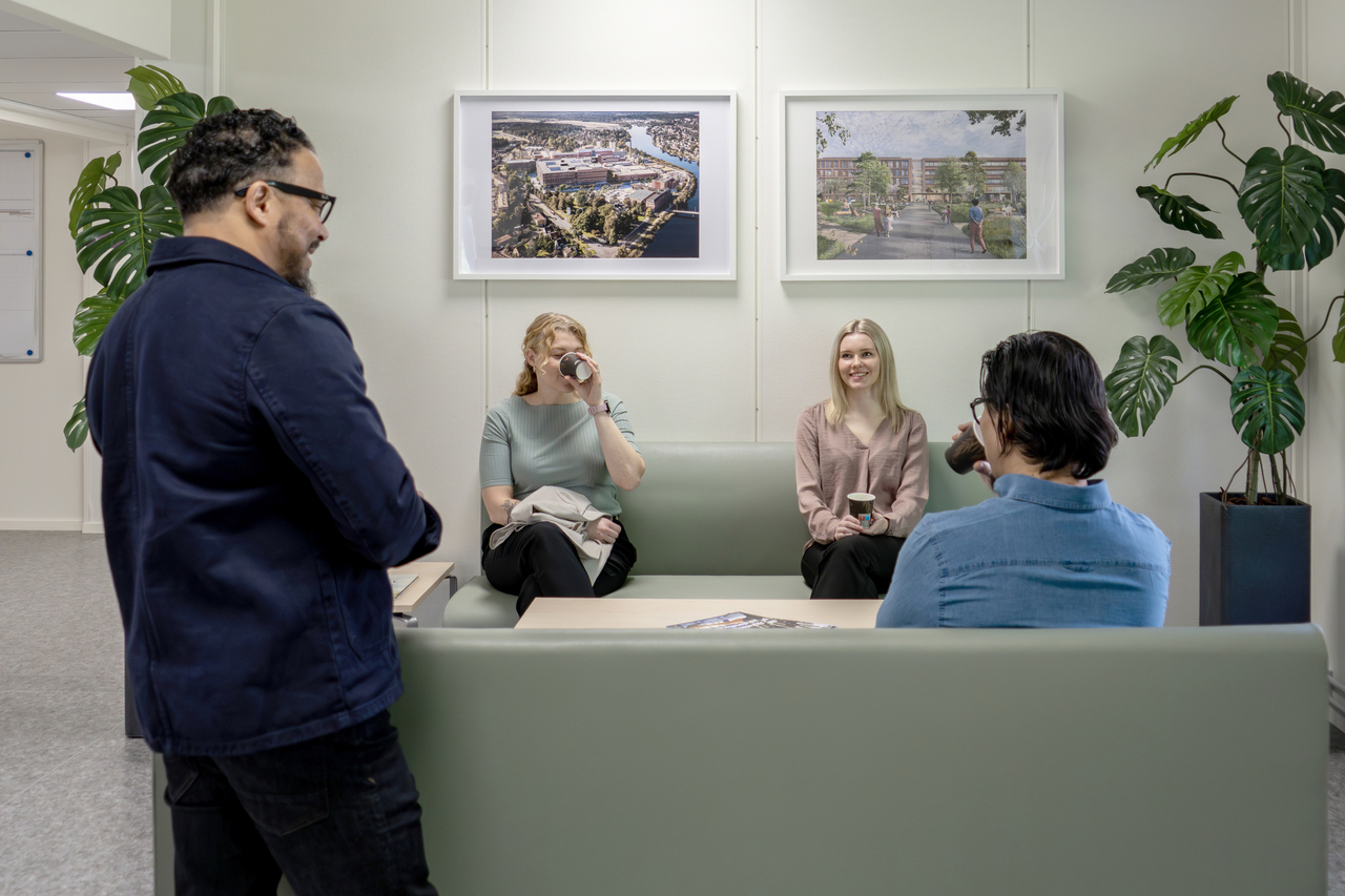 Paus- och mötesyta i kontorsmoduler från Expandia i Skanskas projektkontor i Karlstad. Fyra personer sitter och står runt en soffgrupp i samtal, med kaffekoppar i handen. Väggarna är dekorerade med bilder och gröna växter som skapar en trivsam arbetsmiljö.