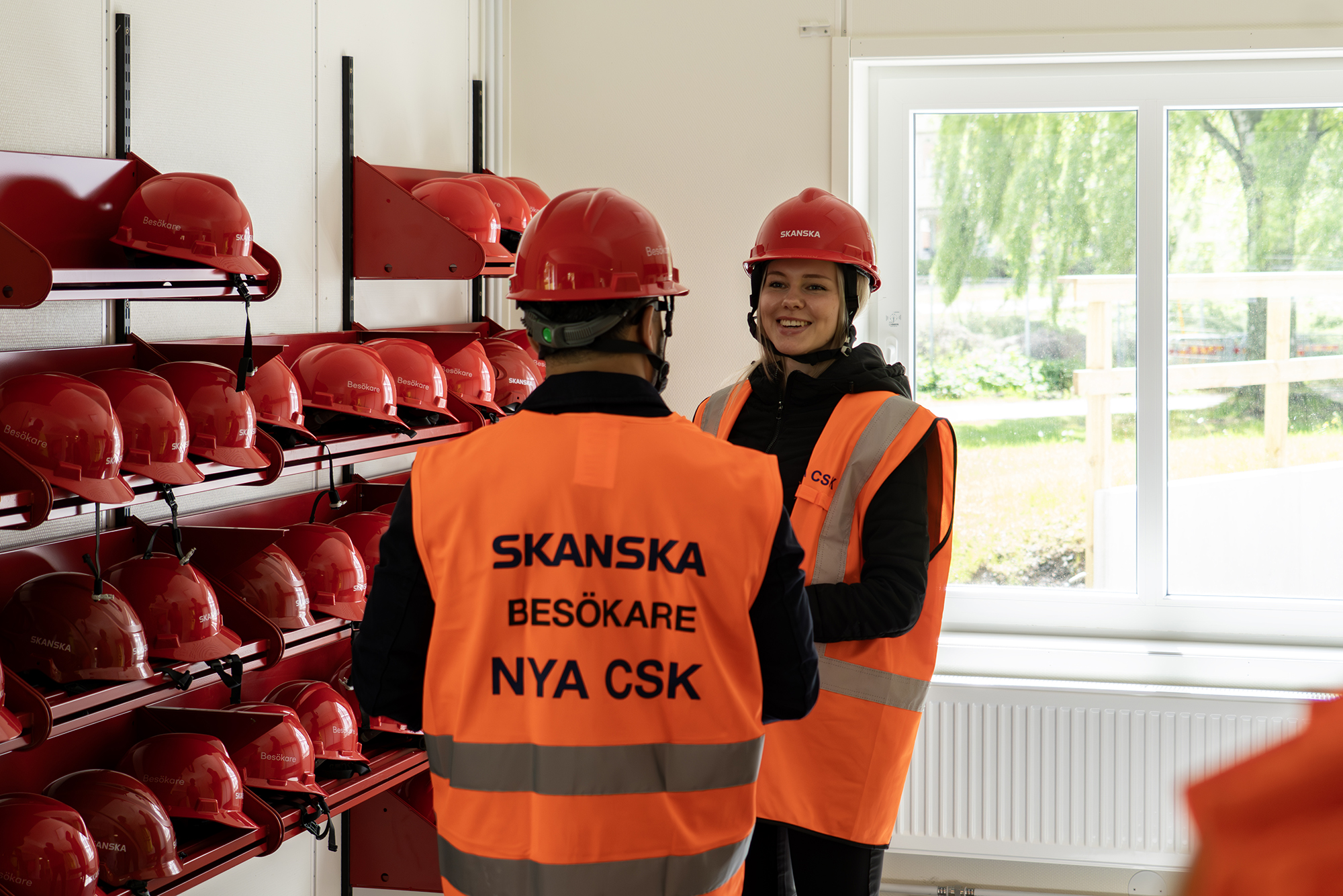 Två personer i varselvästar och röda skyddshjälmar samtalar i ett utrymme för besöksutrustning i Skanskas projektkontor i Karlstad. Rummet har hyllor med märkta skyddshjälmar och ett fönster som släpper in dagsljus, och är placerat i kontorsmoduler från Expandia.