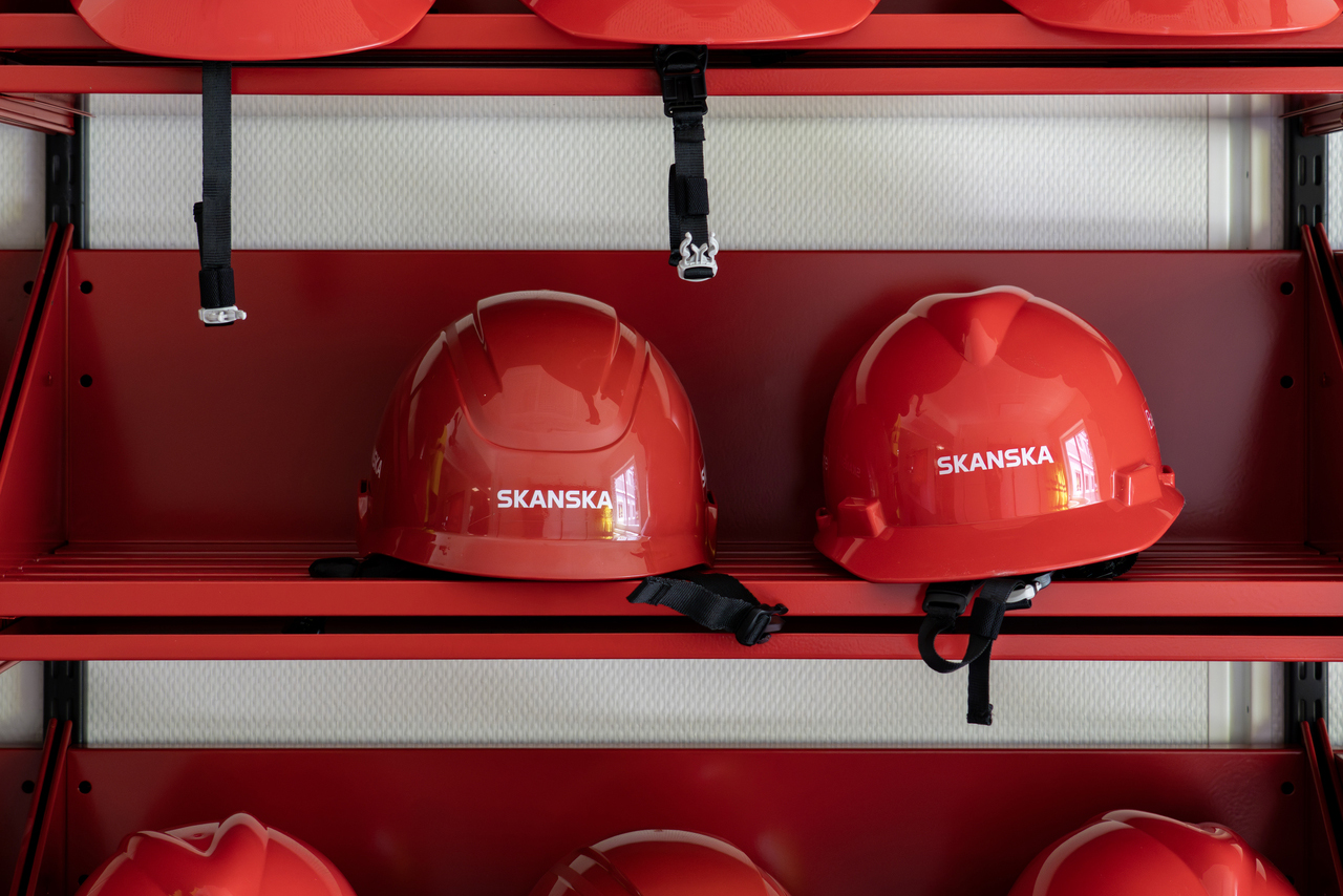 Närbild av röda skyddshjälmar med Skanskas logotyp placerade på ett rött väggställ. Hjälmarna är utrustade med hakband och förvaras prydligt i ett utrymme för besöks- och säkerhetsutrustning i Skanskas projektkontor i Karlstad.