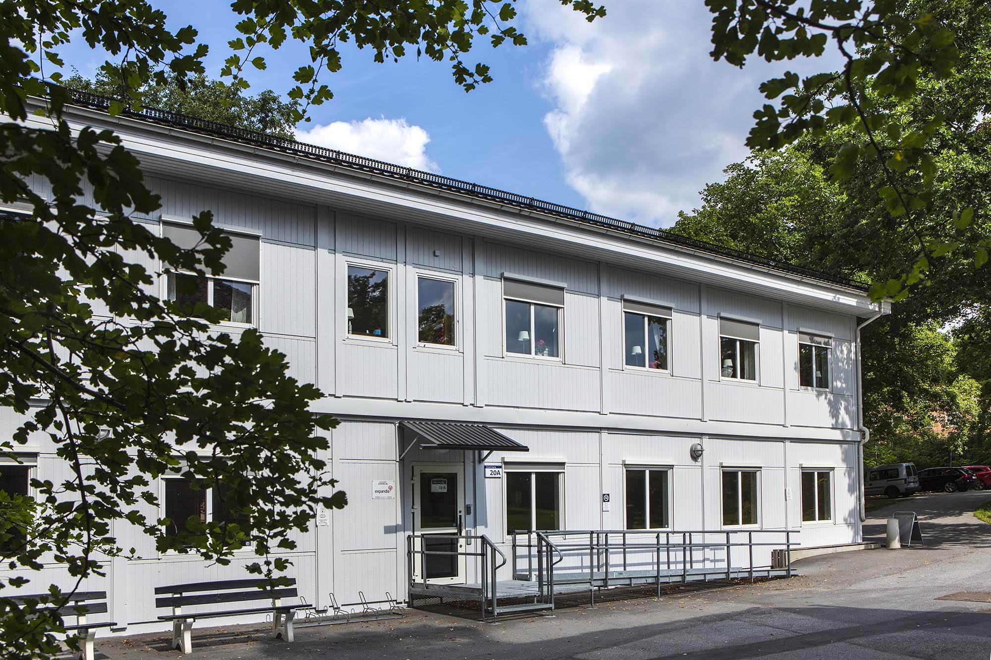 Ljusa kontorsmoduler i två plan med fönsterrader, entré med ramp och räcke, omgivna av grönska och träd i en lugn utemiljö – kontorsmoduler från Expandia.