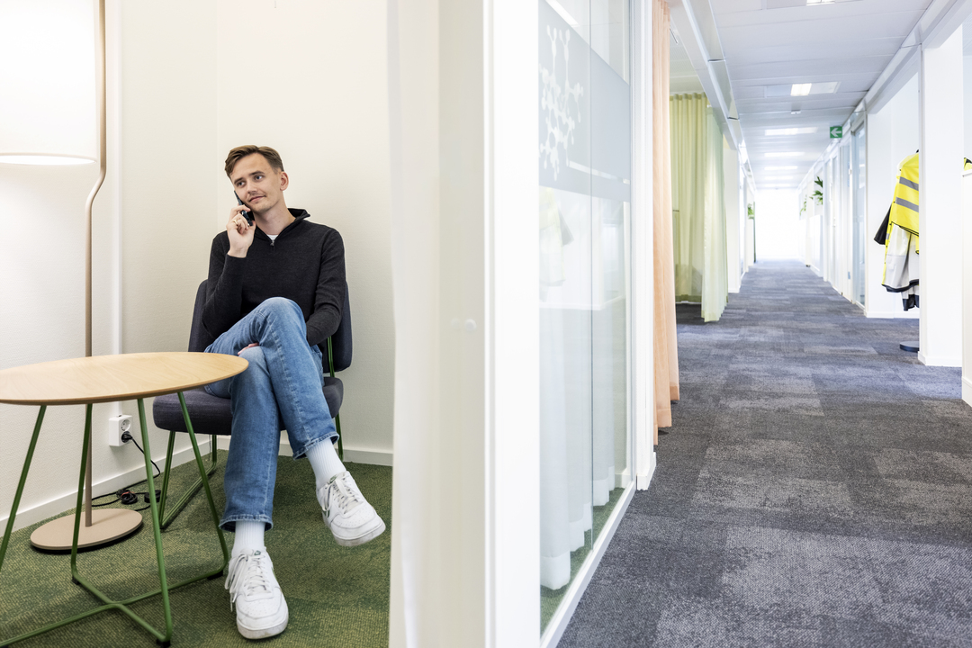Avskild sittplats i kontorsmoduler hos Polypeptide där en medarbetare sitter och pratar i telefon vid ett litet bord, intill glasad korridor med textilgolv och mjuk belysning i en lugn och modern arbetsmiljö – kontorsmoduler från Expandia.