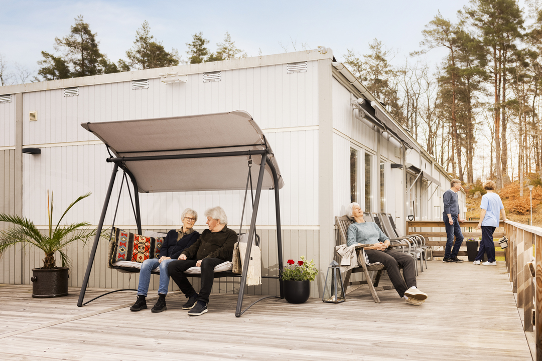 Boende vistas på en uteplats vid Borggården demensvård i Olofström, där några sitter i en hammock och andra vilar på stolar längs trädäcket. I bakgrunden syns en modullösning från Expandia som omger utemiljön.