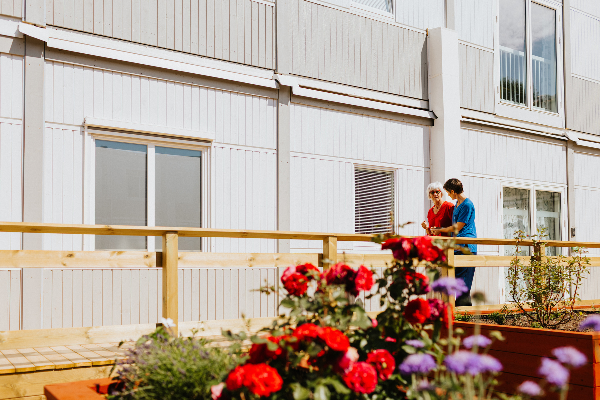 Två personer promenerar och samtalar på en träbelagd gång utanför Herrhagsgården vård- och omsorgsboende i Falun. I förgrunden syns blommande planteringar och i bakgrunden en ljus modullösning från Expandia.