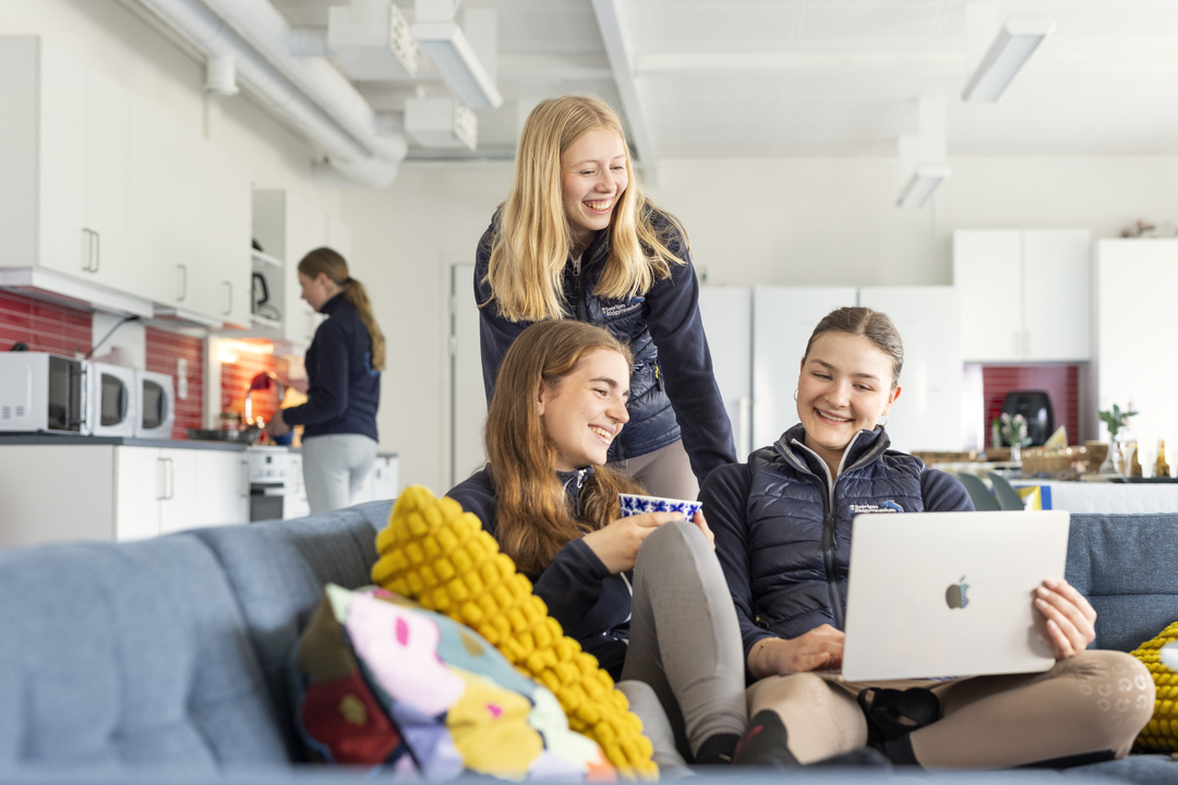Elever vid Sveriges Ridgymnasium i Svedala sitter tillsammans i ett gemensamt inomhusutrymme och samtalar, varav en använder en dator. I bakgrunden syns ett gemensamt kök som ingår i elevboendet.