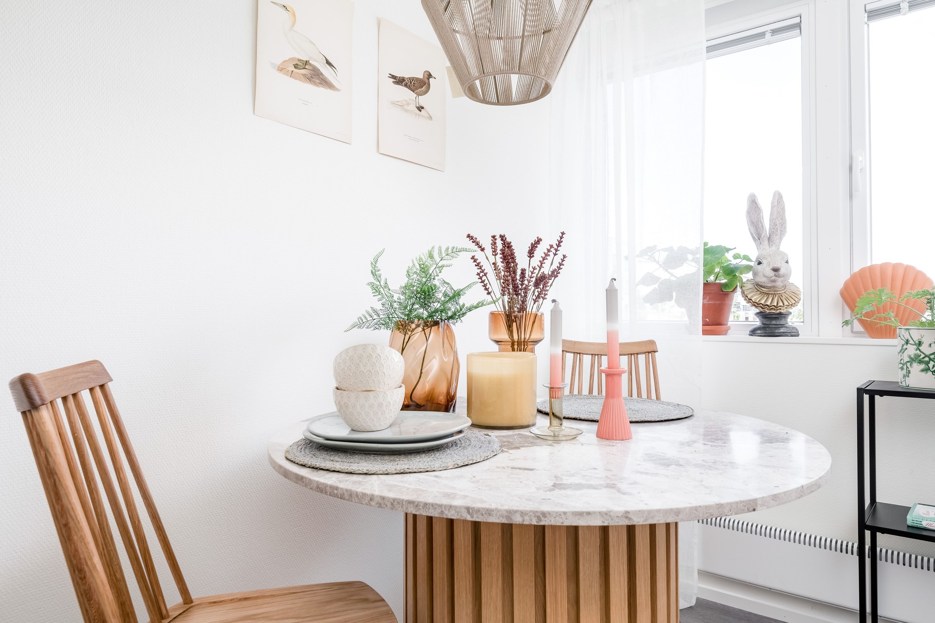 Möblerad studentbostad i Visby med rund matplats i trä, två stolar, dukning och dekorativa detaljer som växter, ljusstakar och konst på väggen, i en ljus och hemtrevlig miljö med dagsljus från fönster, där boendet är uppfört i modulbaserade studentbostäder från Expandia.